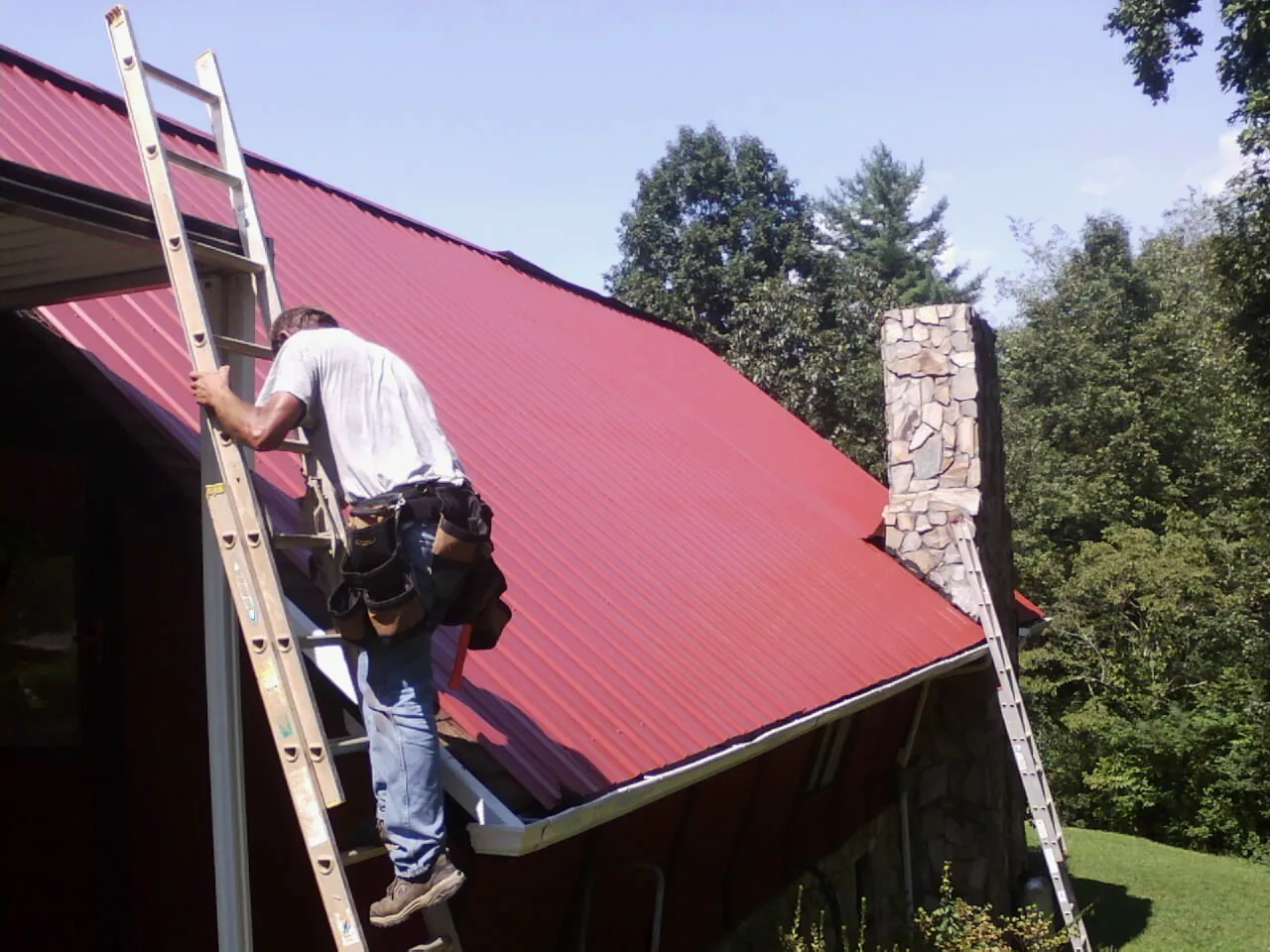 Roofer on a ladder beside a red metal roof and stone chimney — Soffit & Fascia Repair by Roofing Brothers in Duson